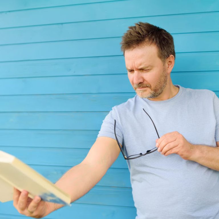 Portrait of mature man with big black eye glasses trying to read book but having difficulties seeing text because of vision problems. Hyperopia, presbyopia.
